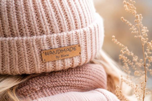 Rechthoekig lichtbruin kunstleren label "Handmade by Bine" op een roze gebreide muts.